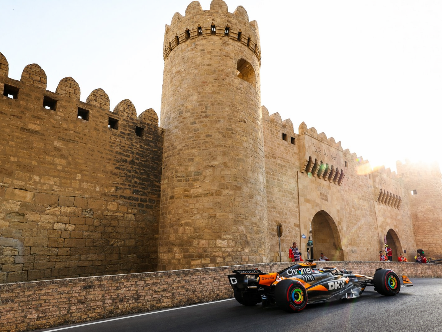 Oranssi-musta Formula 1 -auto ajaa muinaisen kivisen linnoitusmuurin ohi kirkkaassa auringonvalossa.