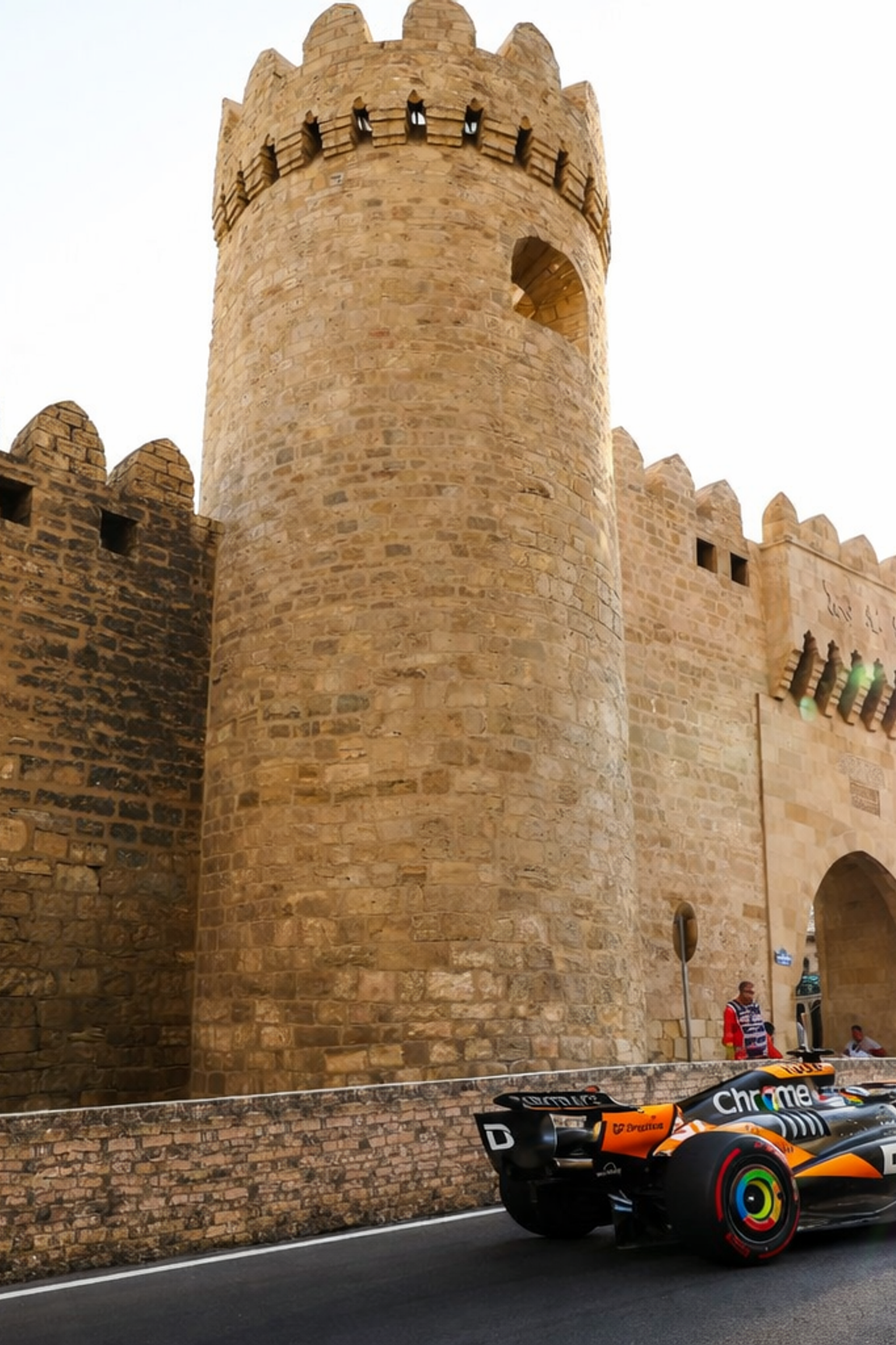 Oranssi-musta Formula 1 -auto ajaa muinaisen kivisen linnoitusmuurin ohi kirkkaassa auringonvalossa.