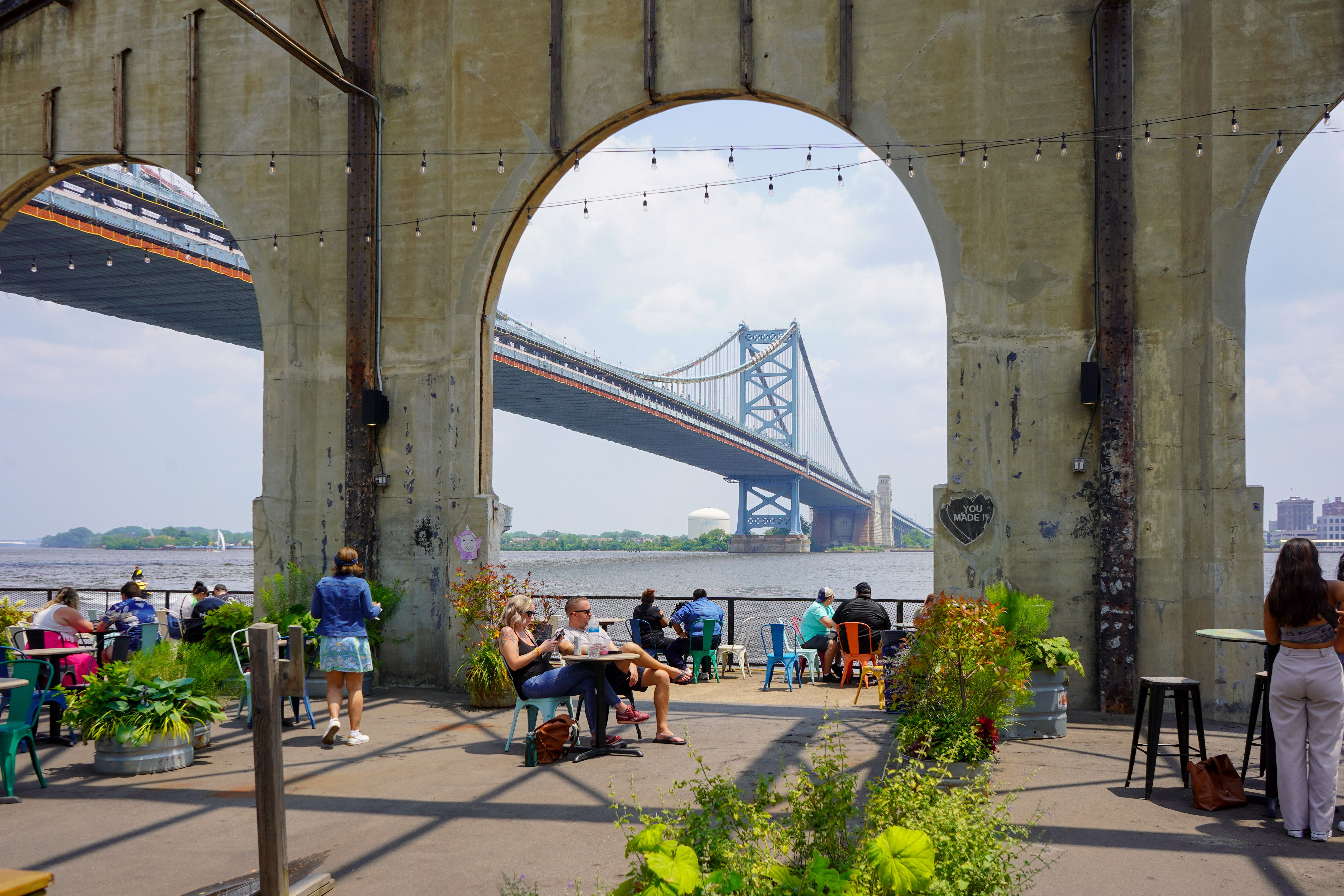 Udendørs spiseområde under betonbuer, med en hængebro over en flod i det fjerne. Philadelphia