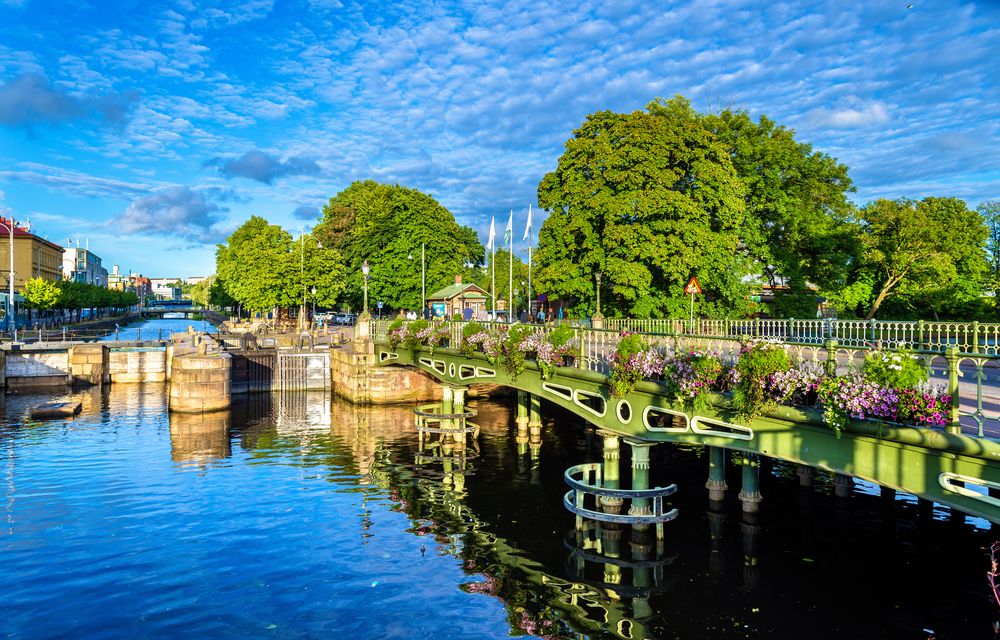 En malerisk kanal med en grøn bro udsmykket med blomster, flankeret af frodige træer, under en klar blå, skyet himmel.