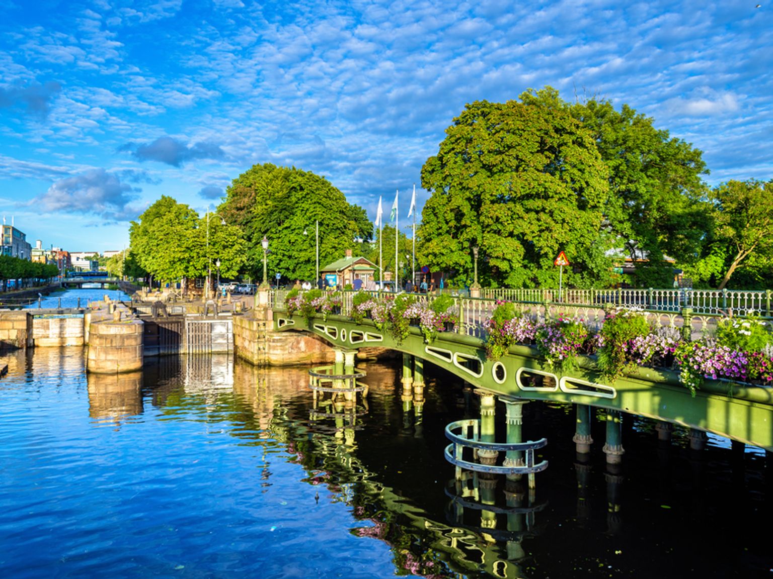 En malerisk kanal med en grøn bro udsmykket med blomster, flankeret af frodige træer, under en klar blå, skyet himmel.
