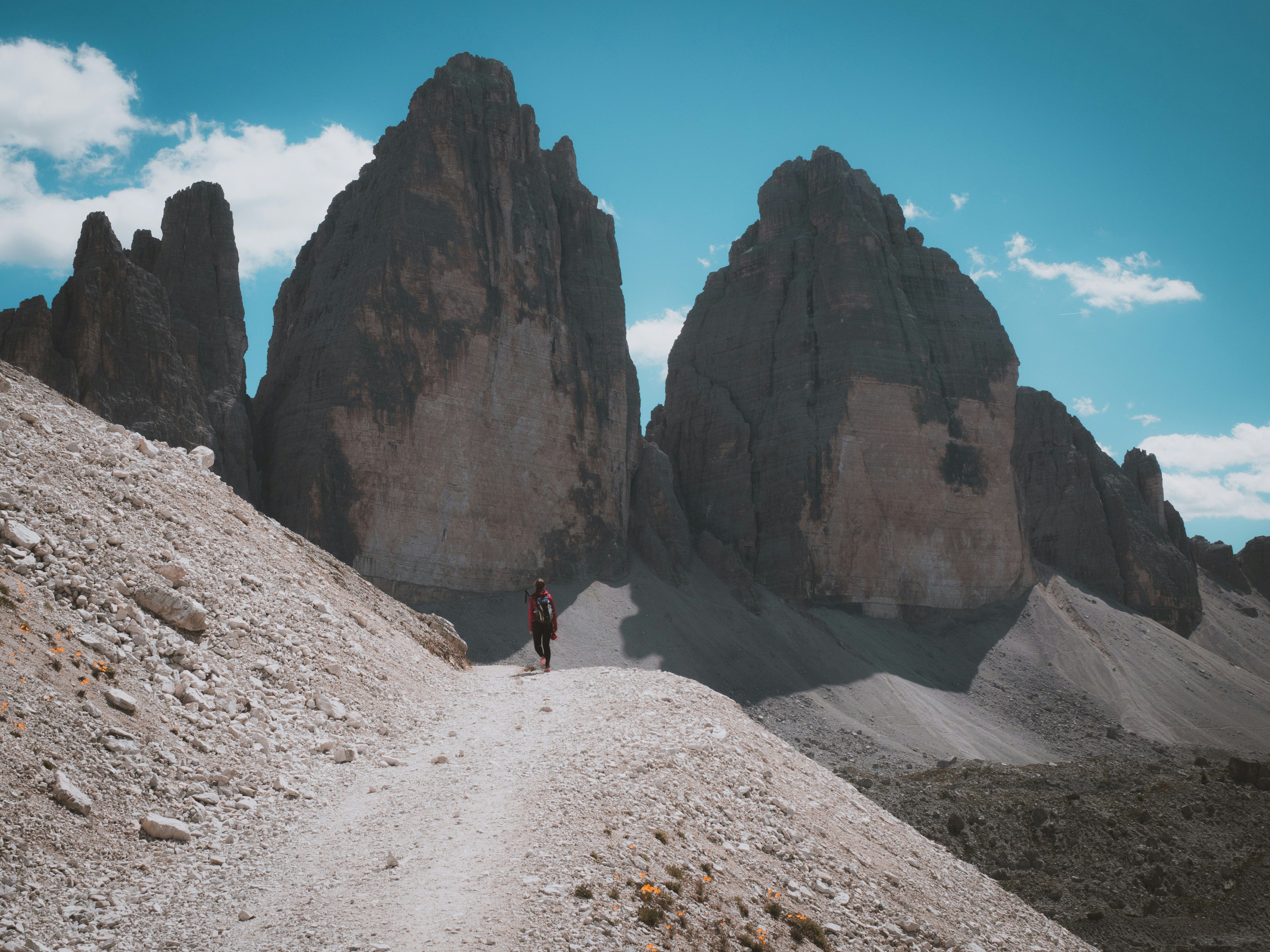 En turgåer går på en steinete sti med fjellene Tre Cime di Lavaredo i bakgrunnen under en blå himmel.
