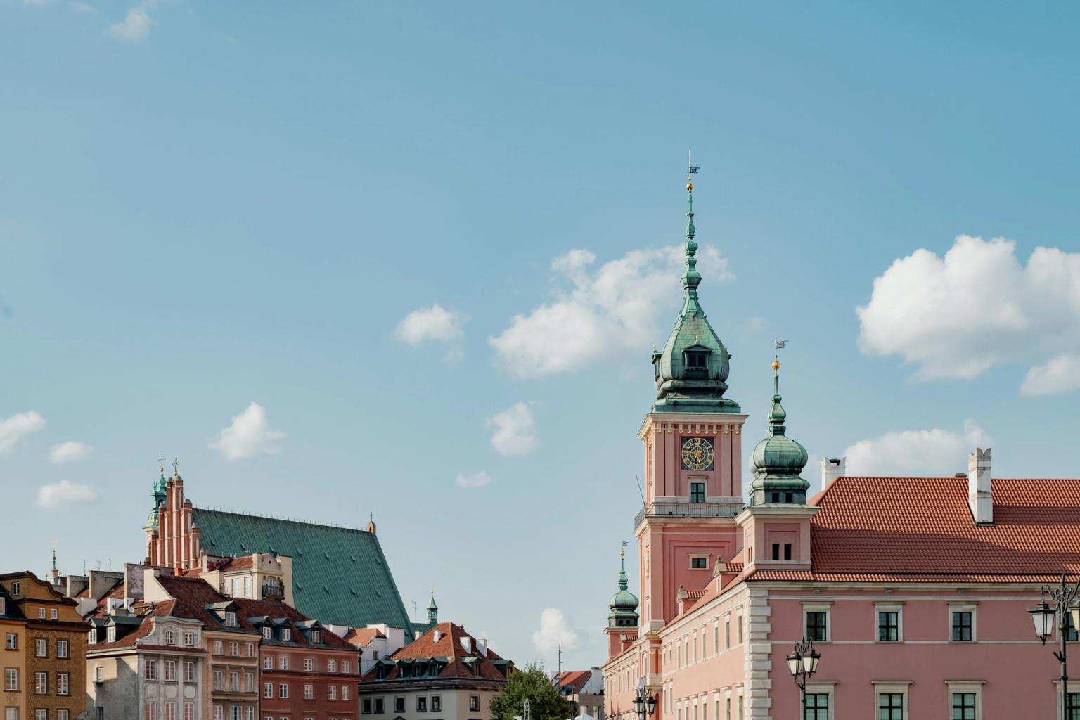 Warszawas gamla stads silhuett med det rosa kungliga slottet och en katedral med grönt tak under en delvis molnig himmel.