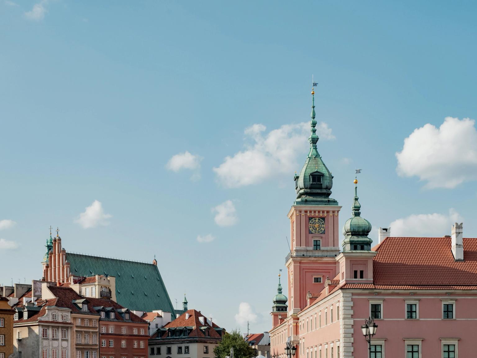 Warszawas gamla stads silhuett med det rosa kungliga slottet och en katedral med grönt tak under en delvis molnig himmel.