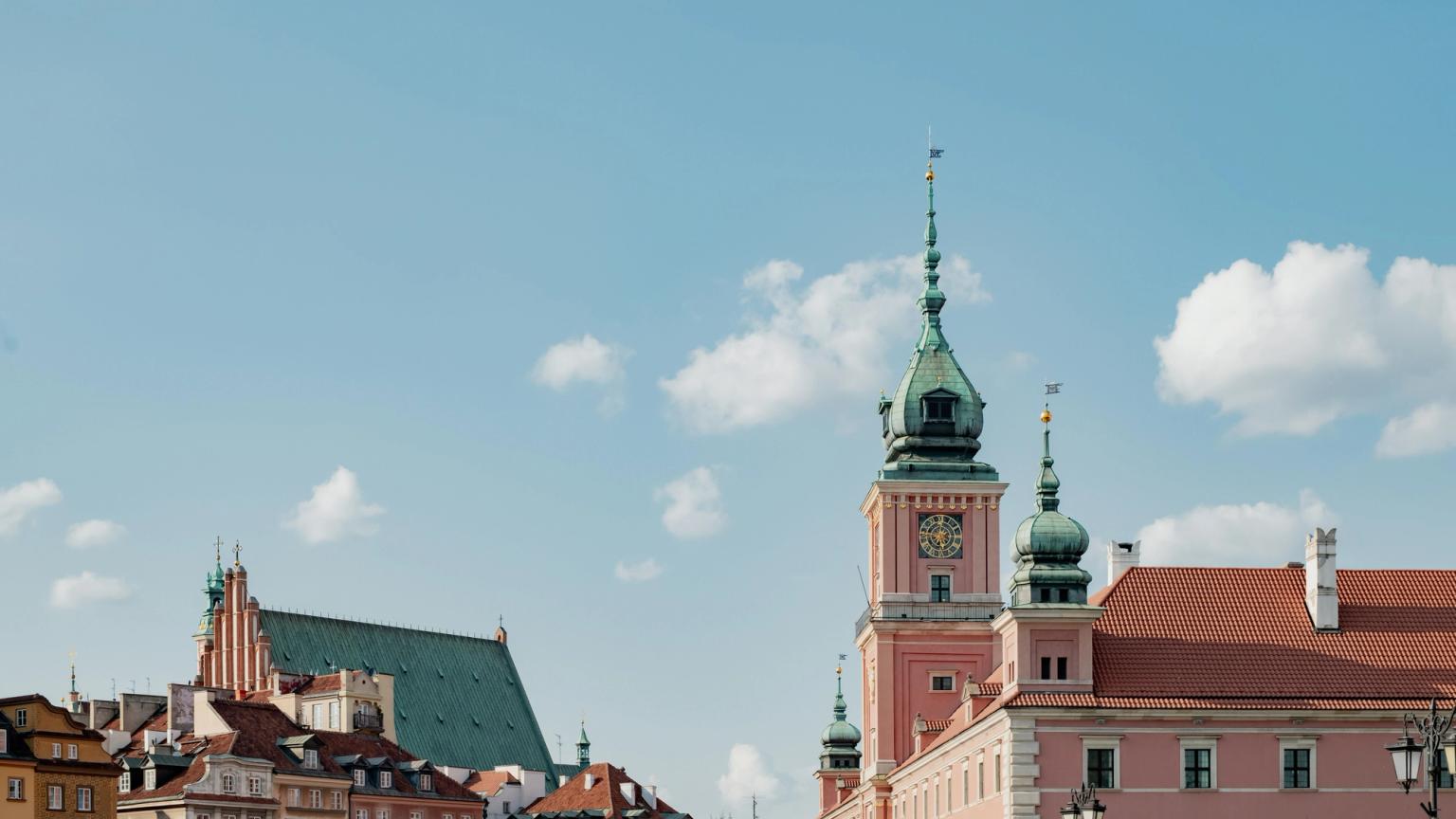 Warszawas gamla stads silhuett med det rosa kungliga slottet och en katedral med grönt tak under en delvis molnig himmel.