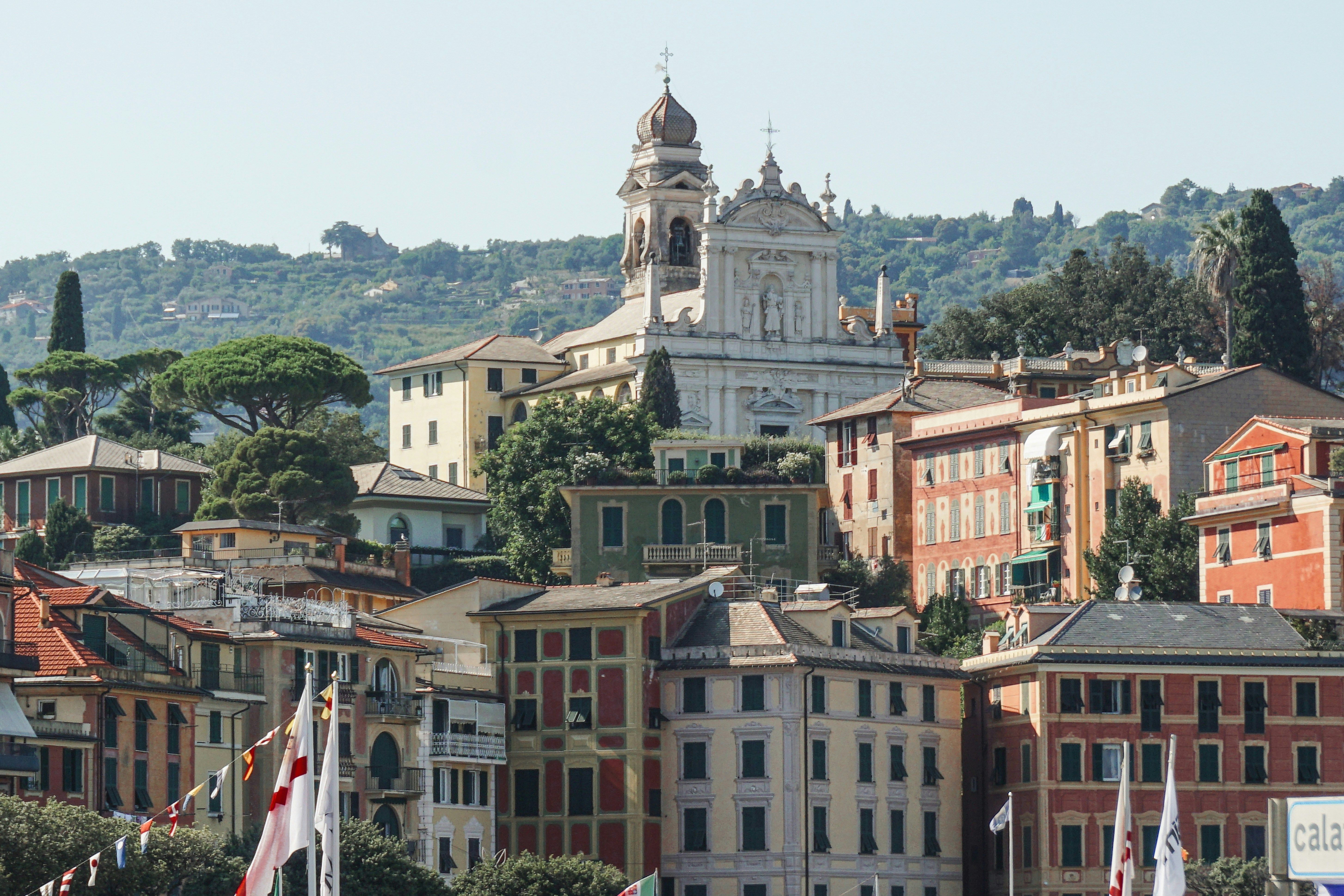 En farverig bjergskråningsby med en fremtrædende hvid kirke og klokketårn, som ligger indlejret op ad et grønt bjerg. Santa Margherita Ligure