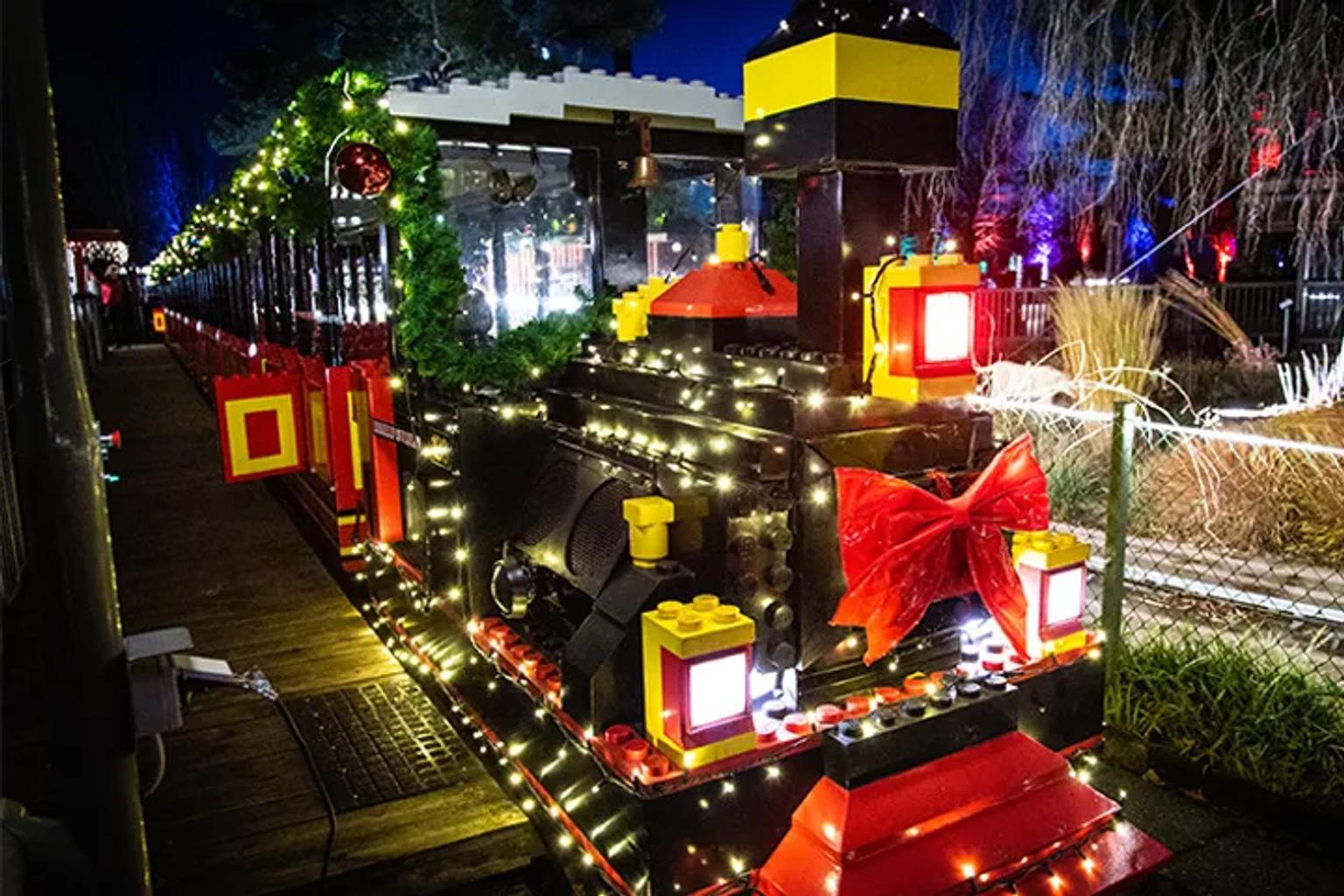 Ett festligt LEGO-tåg täckt med vita julbelysningar och en stor röd rosett på natten. Legoland