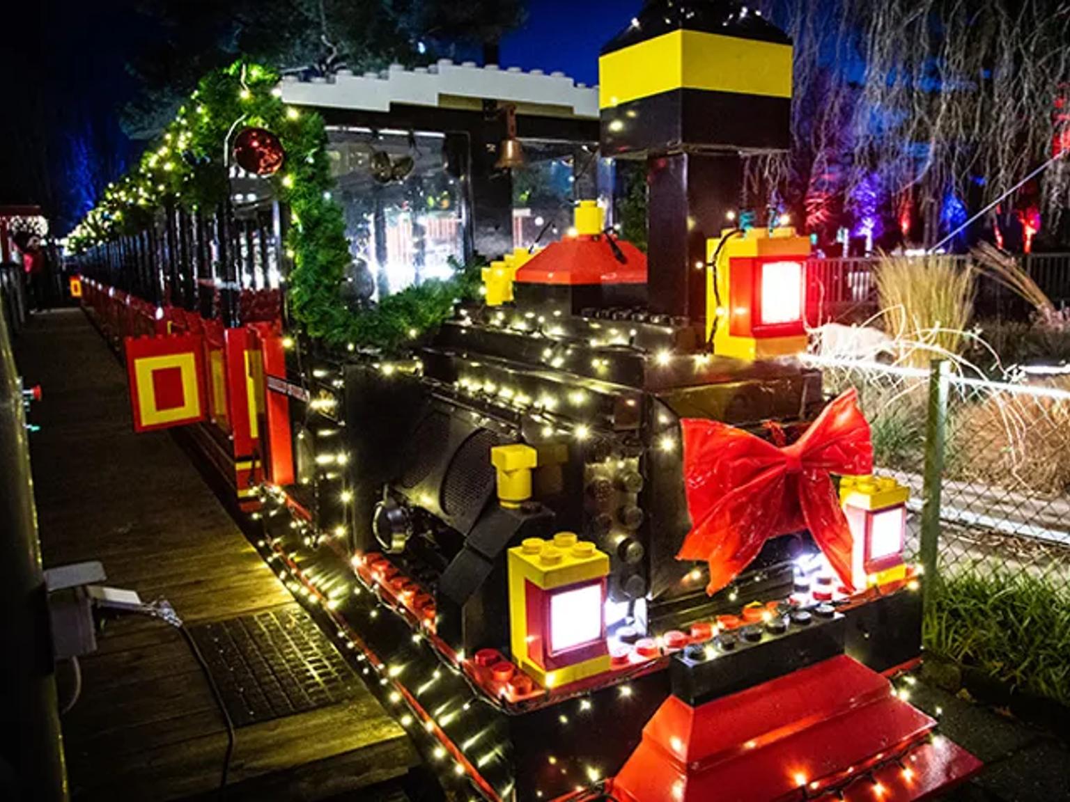 Et festlig LEGO-tog dekket med hvite julelys og en stor rød sløyfe om natten. Legoland