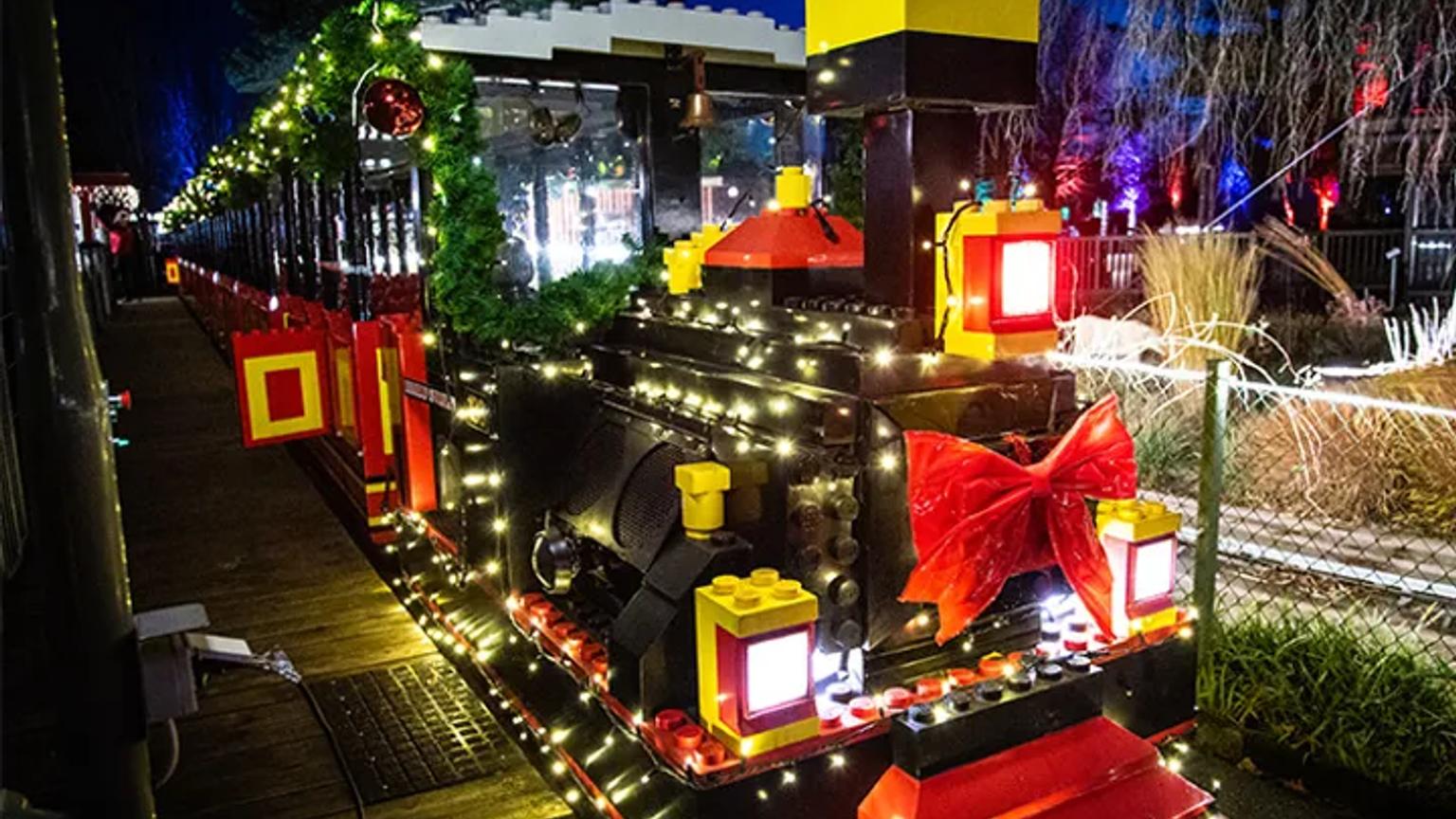 Ett festligt LEGO-tåg täckt med vita julbelysningar och en stor röd rosett på natten. Legoland