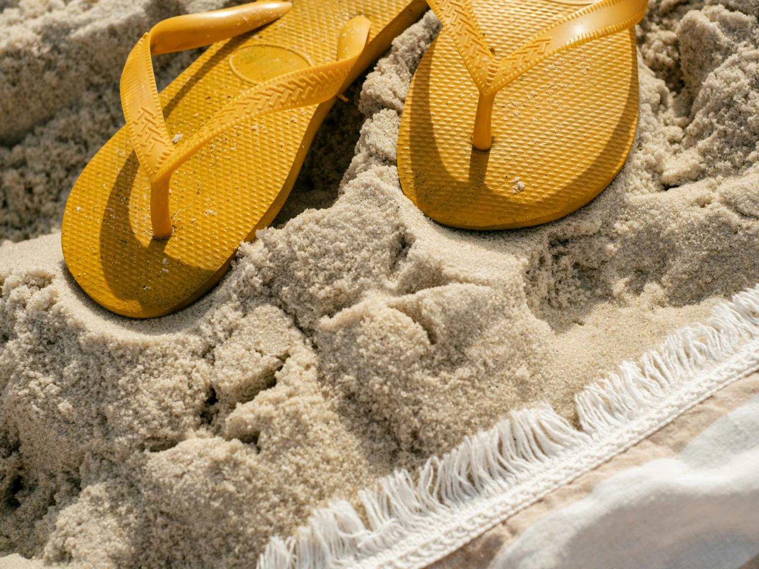 Gula flipflops som vilar i sanden bredvid en fransad handduk.