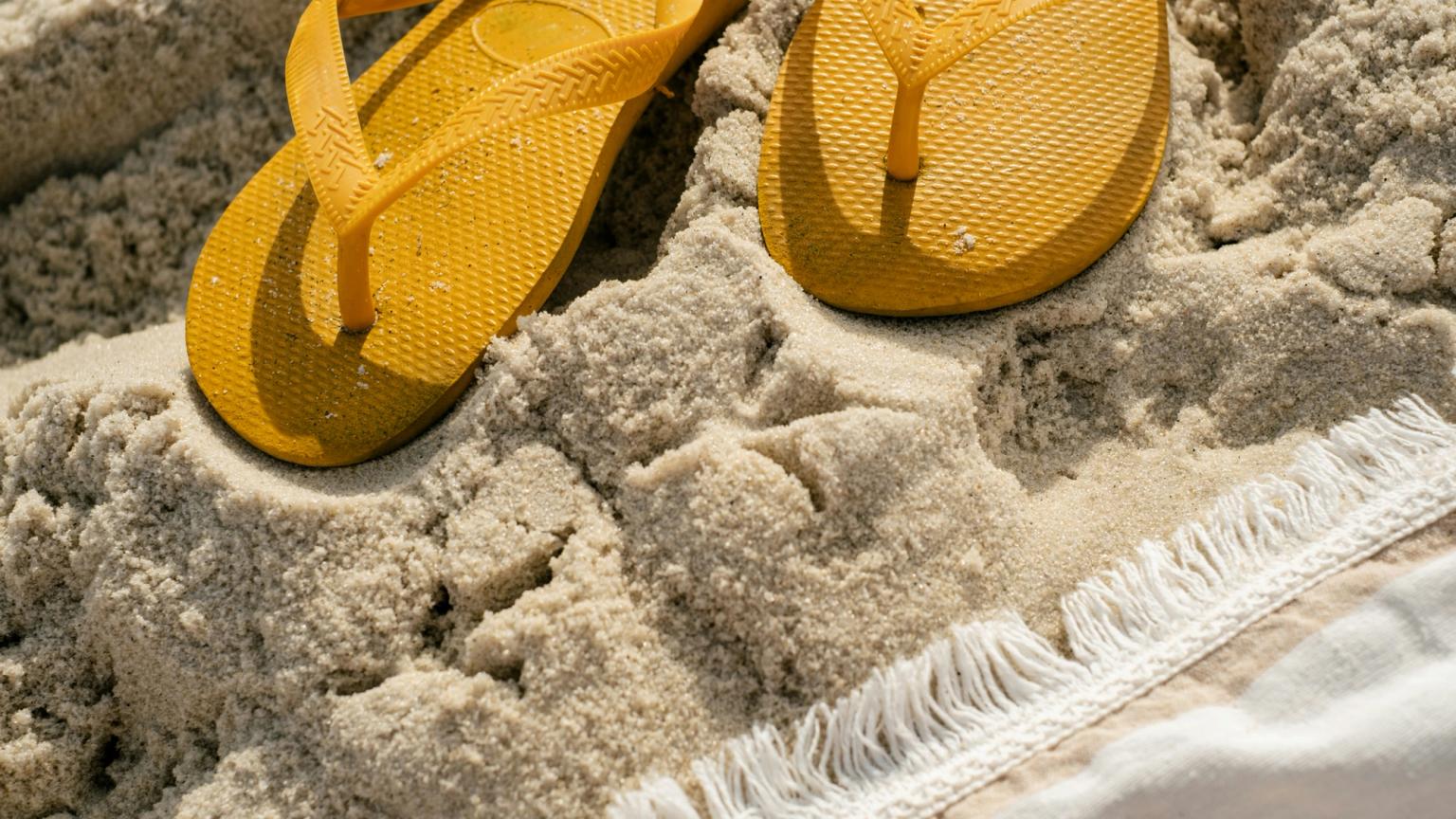 Gula flipflops som vilar i sanden bredvid en fransad handduk.