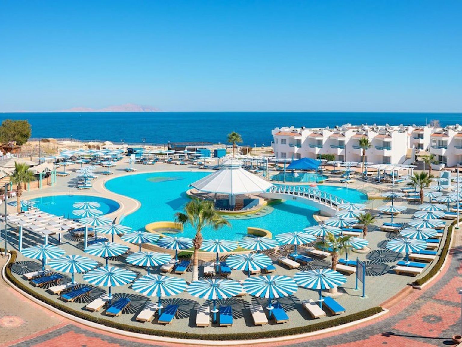 Flygfoto av en solig resort med stora swimmingpooler, blå- och vitrandiga parasoller och resortbyggnader med utsikt över havet.