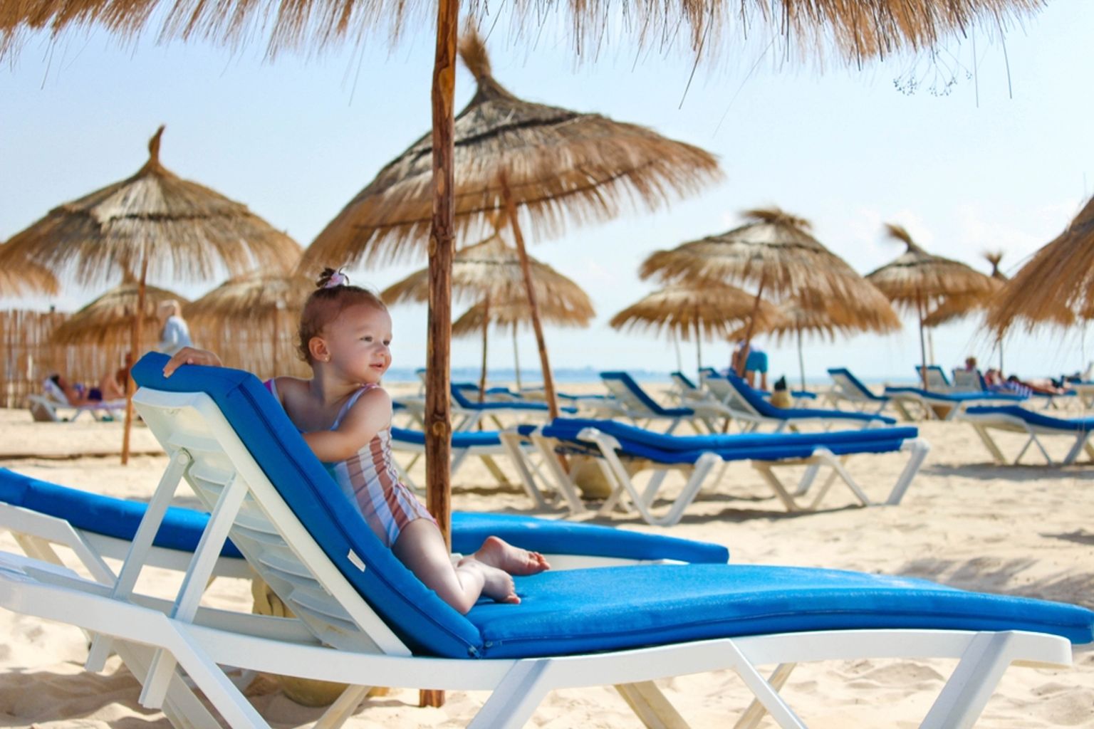 Ett småbarn i en randig baddräkt sitter på en blå strandstol på en solig sandstrand med parasoller av halm i bakgrunden.
