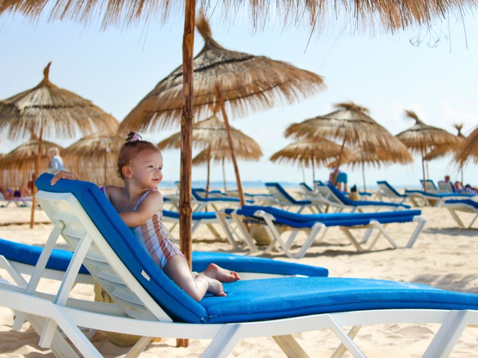 Ett småbarn i en randig baddräkt sitter på en blå strandstol på en solig sandstrand med parasoller av halm i bakgrunden.