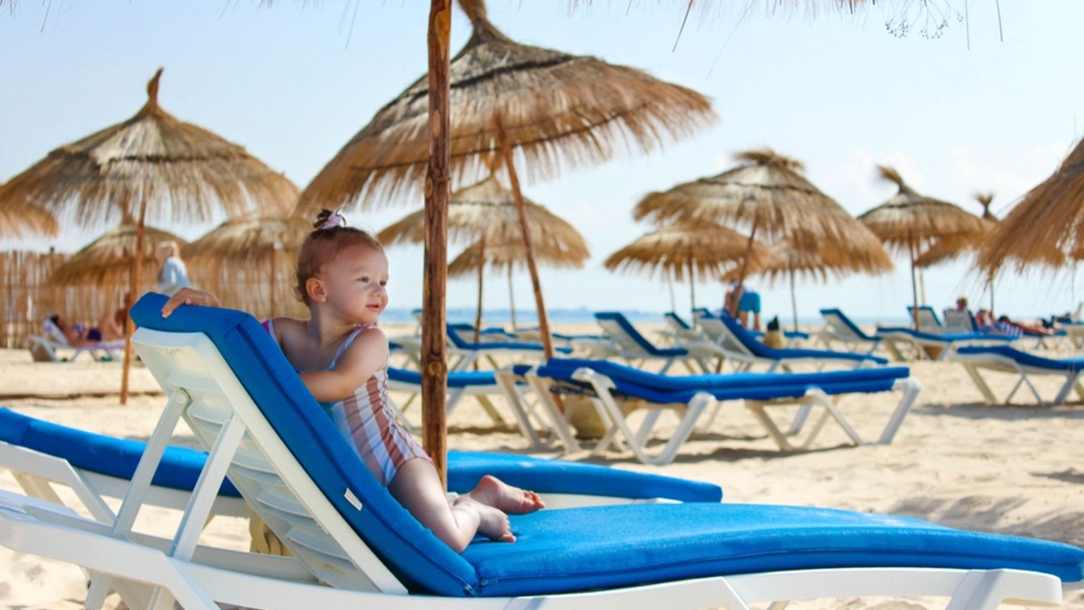 Ett småbarn i en randig baddräkt sitter på en blå strandstol på en solig sandstrand med parasoller av halm i bakgrunden.