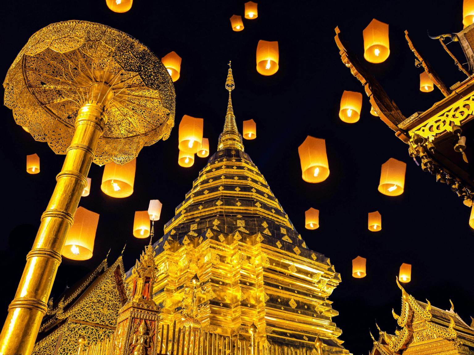Mange lysende himmellykter svever over et gyllent buddhisttempel og seremoniell paraply om natten i Thailand.