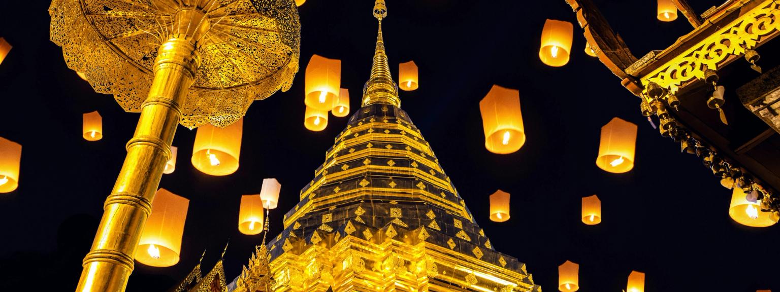 Mange lysende himmellykter svever over et gyllent buddhisttempel og seremoniell paraply om natten i Thailand.