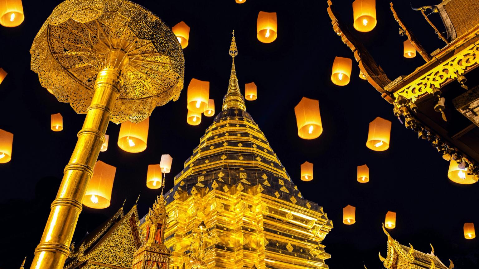 Mange lysende himmellygter svæver over et gyldent buddhisttempel og ceremonielt paraply om natten i Thailand.
