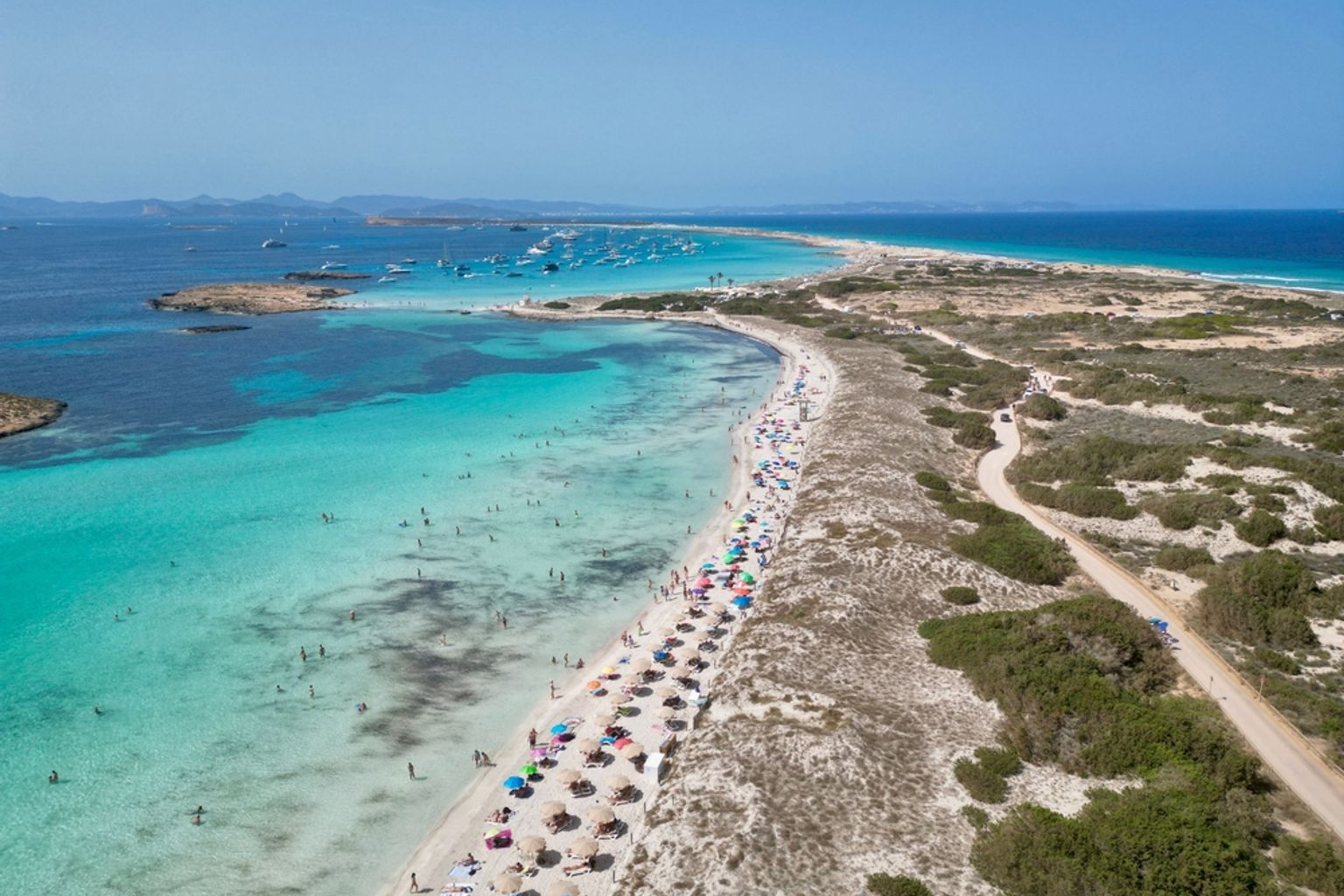 Flygfoto av en livlig strand med turkost vatten, rader av färgglada parasoller och en smal sandremsa som leder till en vik med många båtar. Formentera, Spanien