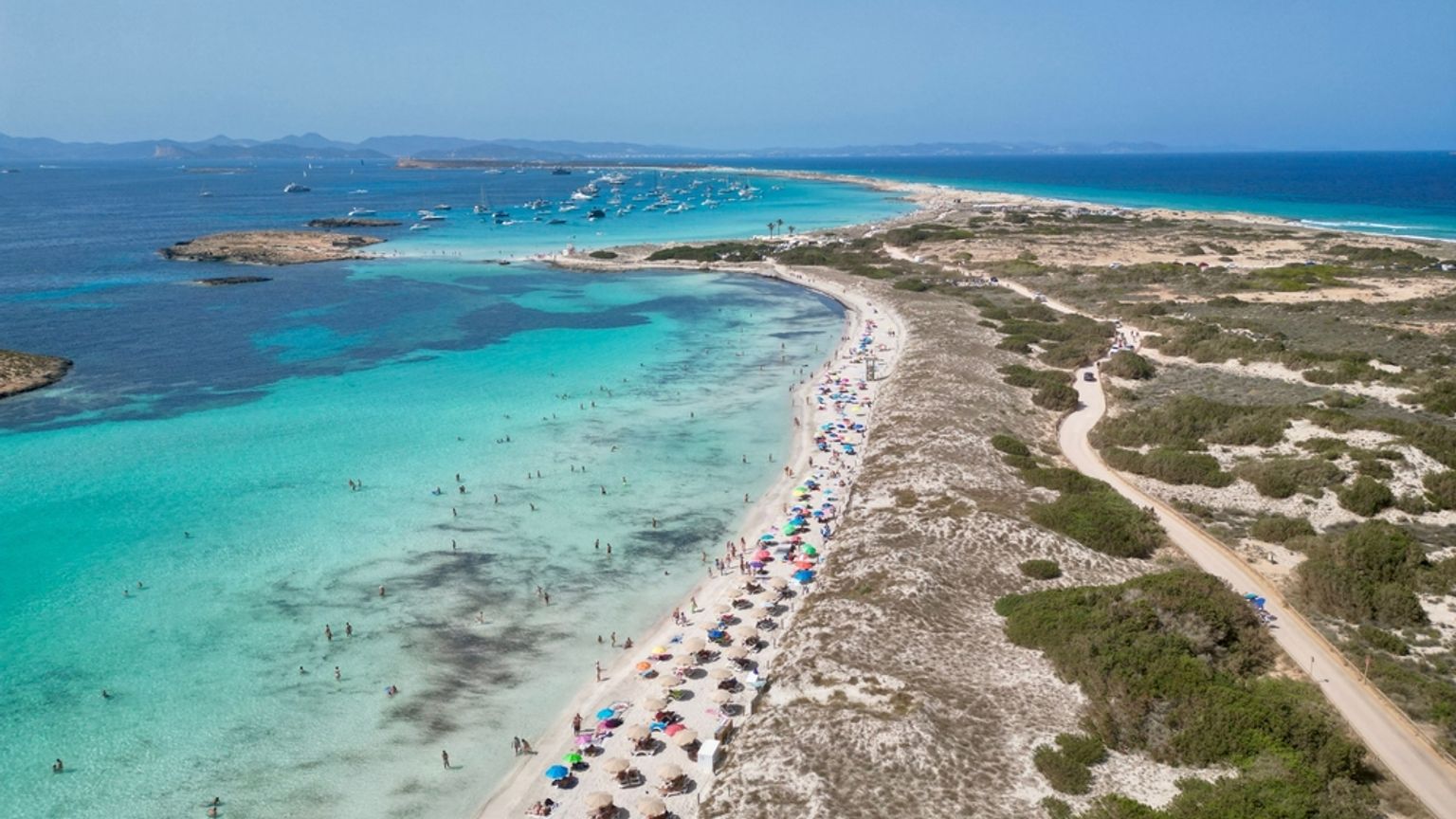 Flygfoto av en livlig strand med turkost vatten, rader av färgglada parasoller och en smal sandremsa som leder till en vik med många båtar. Formentera, Spanien