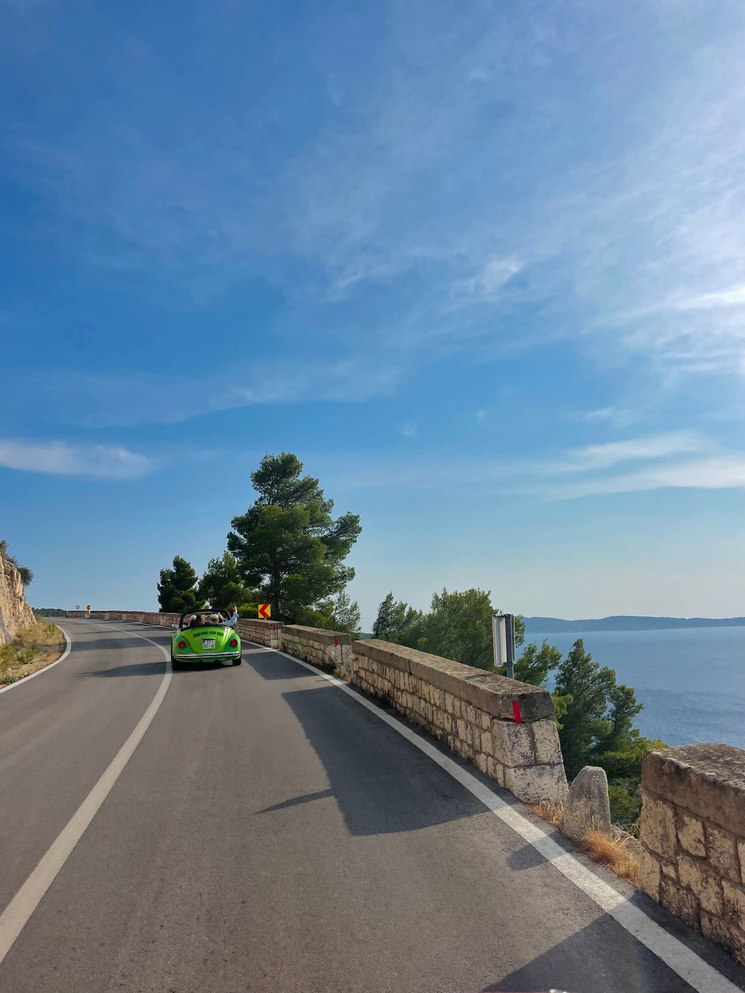 Un cabriolet vert roule sur une route côtière sinueuse sur l’île de Vis en Croatie, sous un ciel bleu clair, avec la mer visible sur la droite.