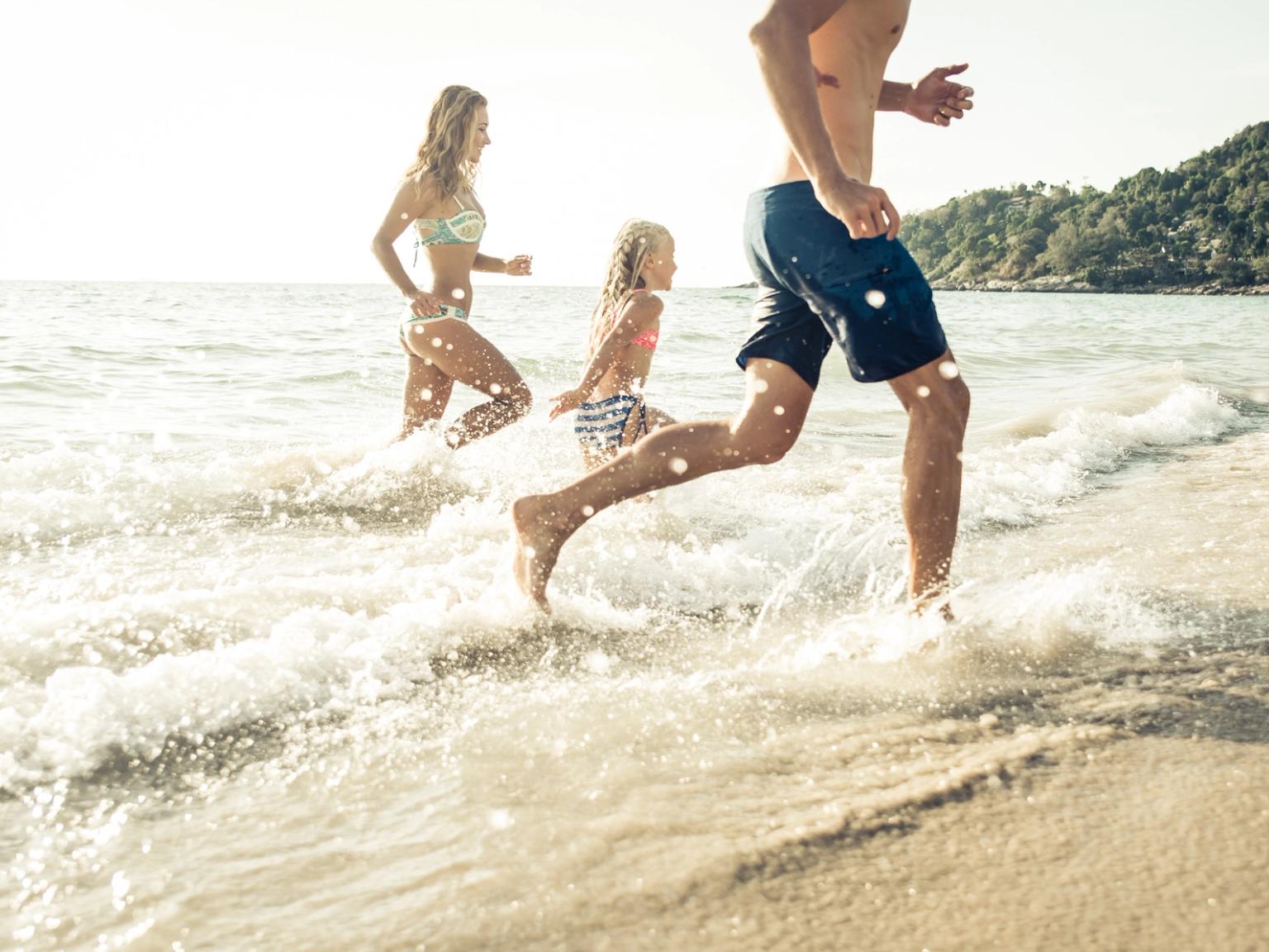 En familj springer genom grunda vågor på en solig strand.