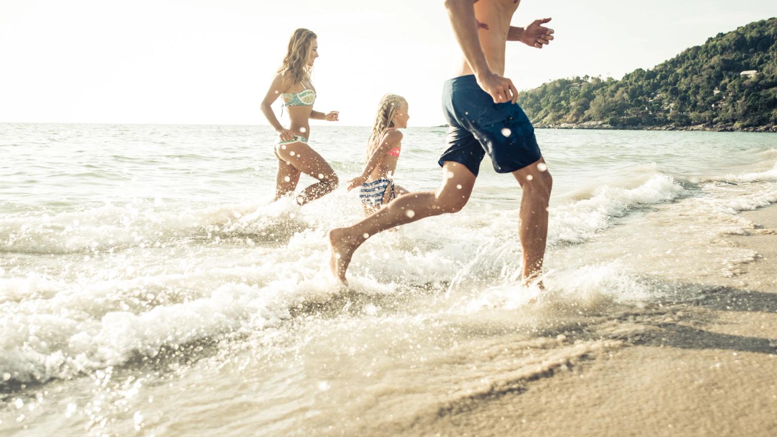 En familj springer genom grunda vågor på en solig strand.