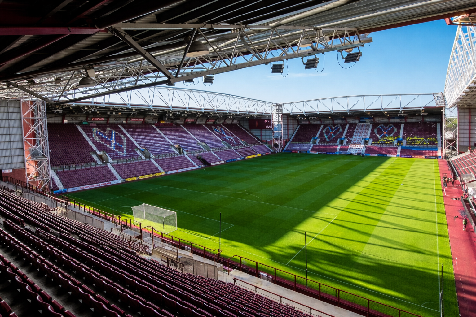 Leeg voetbalstadion met bordeauxrode stoelen die hartvormen vormen op de tribunes, en een groen veld.