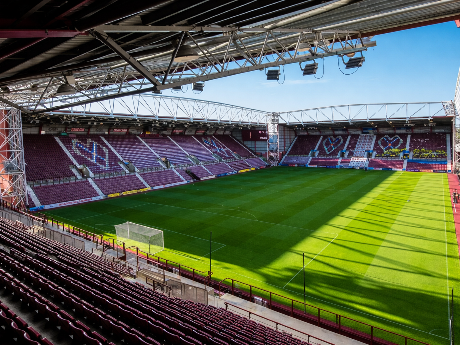 Leeg voetbalstadion met bordeauxrode stoelen die hartvormen vormen op de tribunes, en een groen veld.