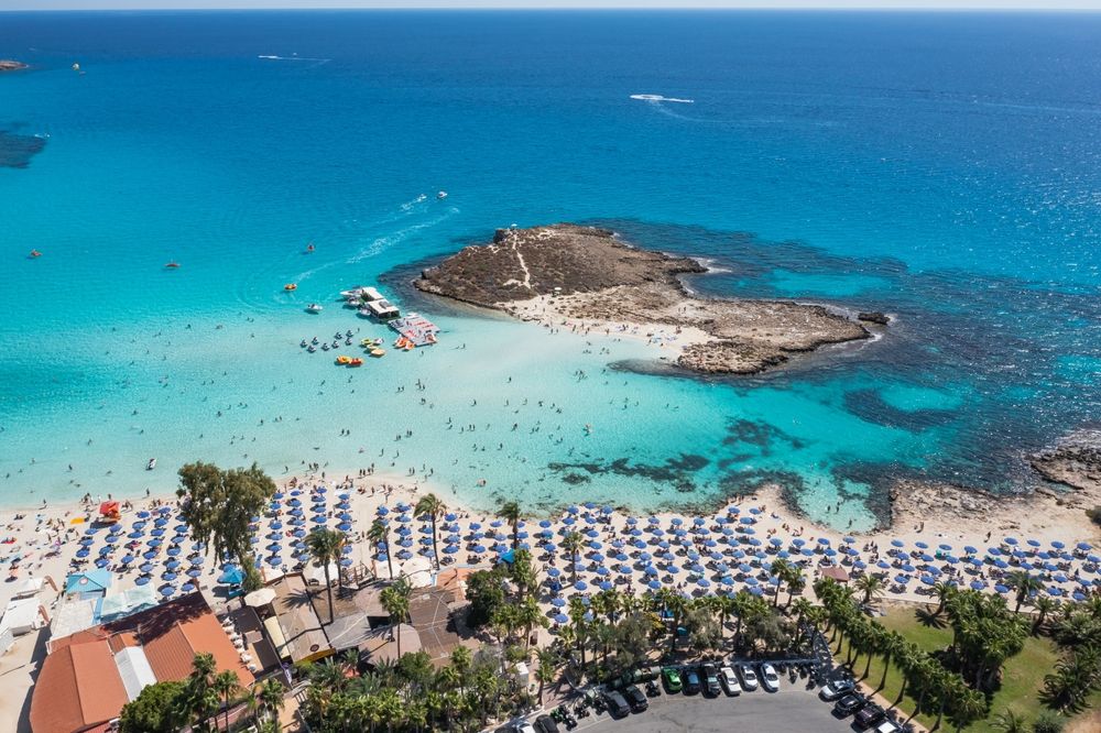 Luftfoto av en fullsatt strand med mange blå parasoller, mennesker som svømmer i turkis vann, og steinete småøyer.