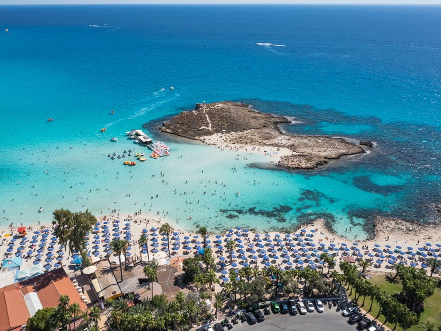 Luftfoto av en fullsatt strand med mange blå parasoller, mennesker som svømmer i turkis vann, og steinete småøyer.