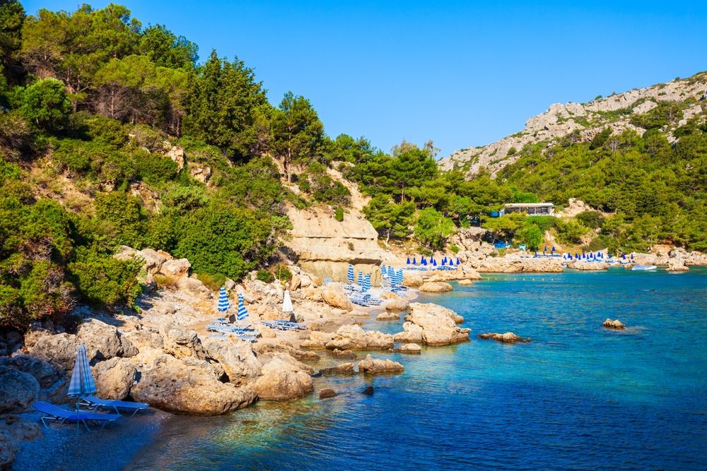 Rocky beach with turquoise water, green hills, and blue beach umbrellas. Anthony Quinn Bay, Rhodes