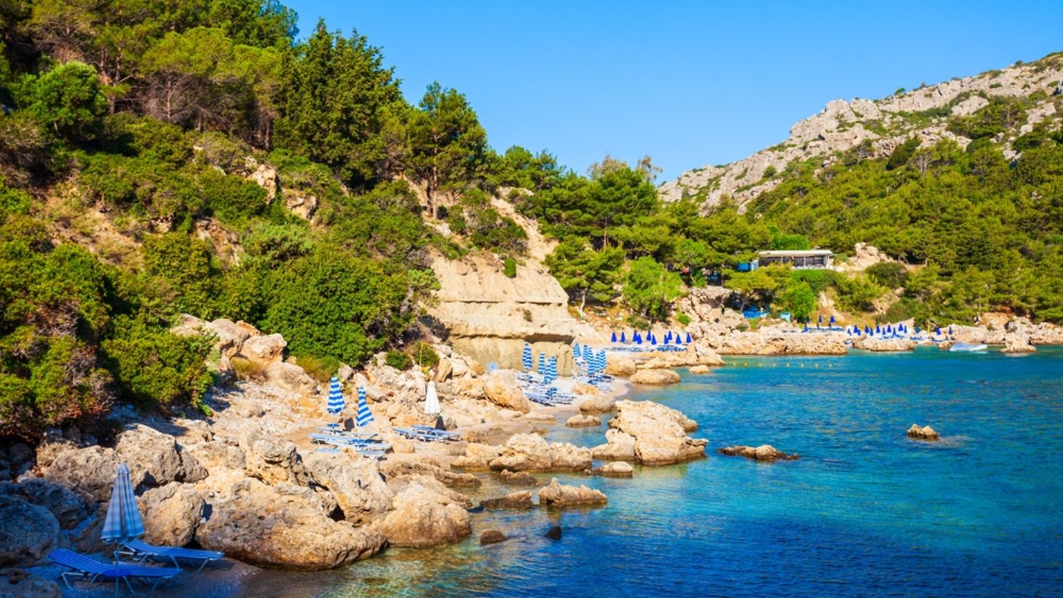 Klippig strand med turkost vatten, gröna kullar och blå strandparasoller. Anthony Quinn Bay, Rhodos