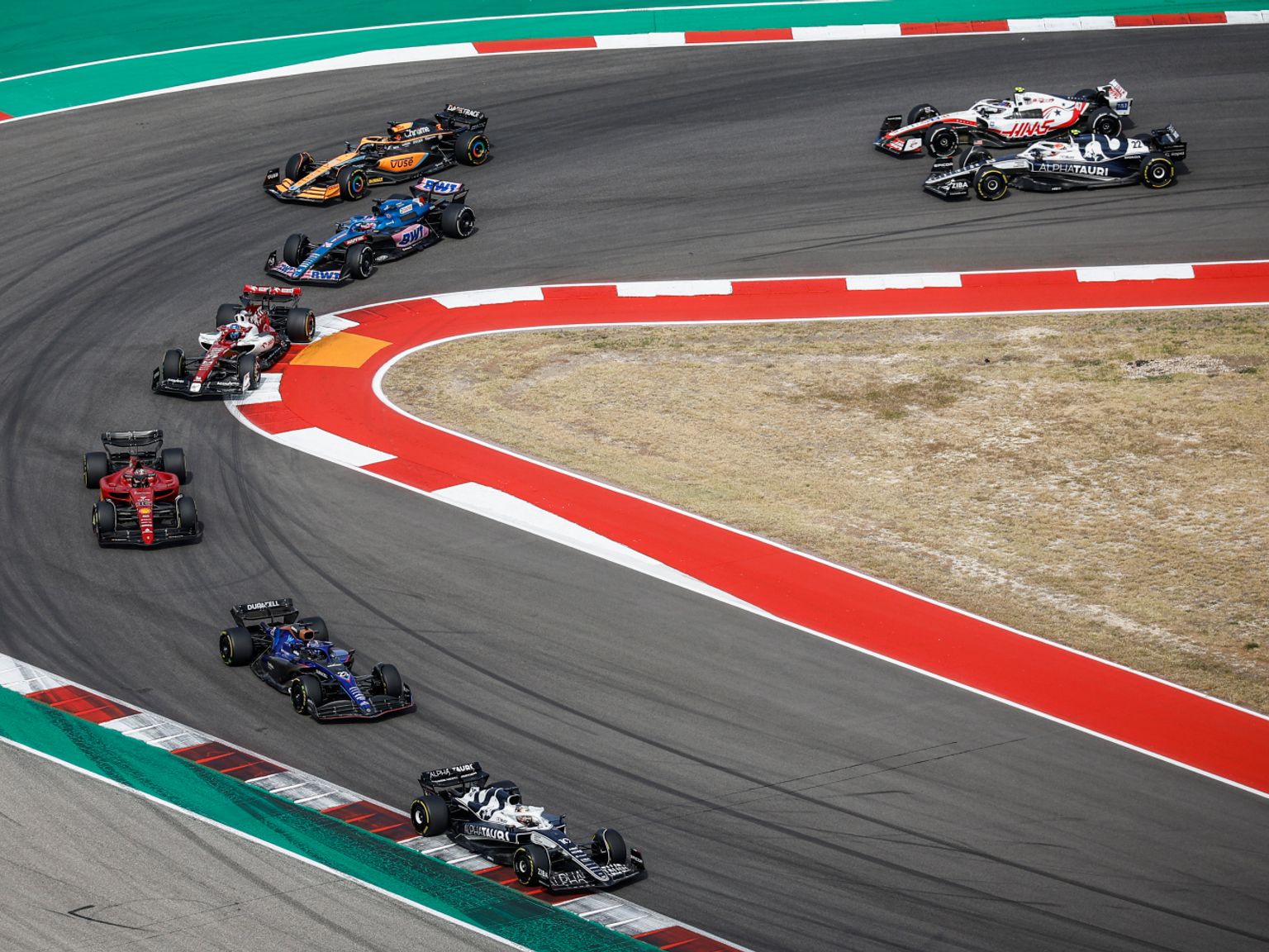 Vários carros de Fórmula 1 navegam em uma curva larga em uma pista de corrida.