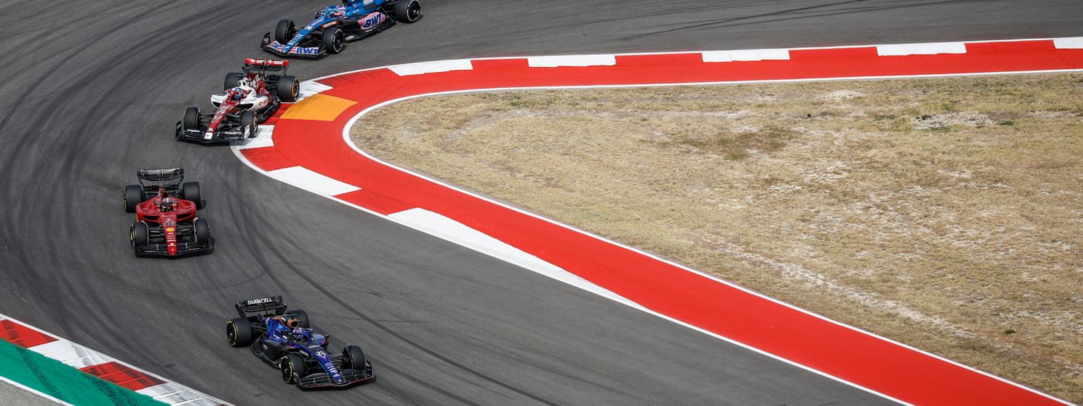 Vários carros de Fórmula 1 navegam em uma curva larga em uma pista de corrida.