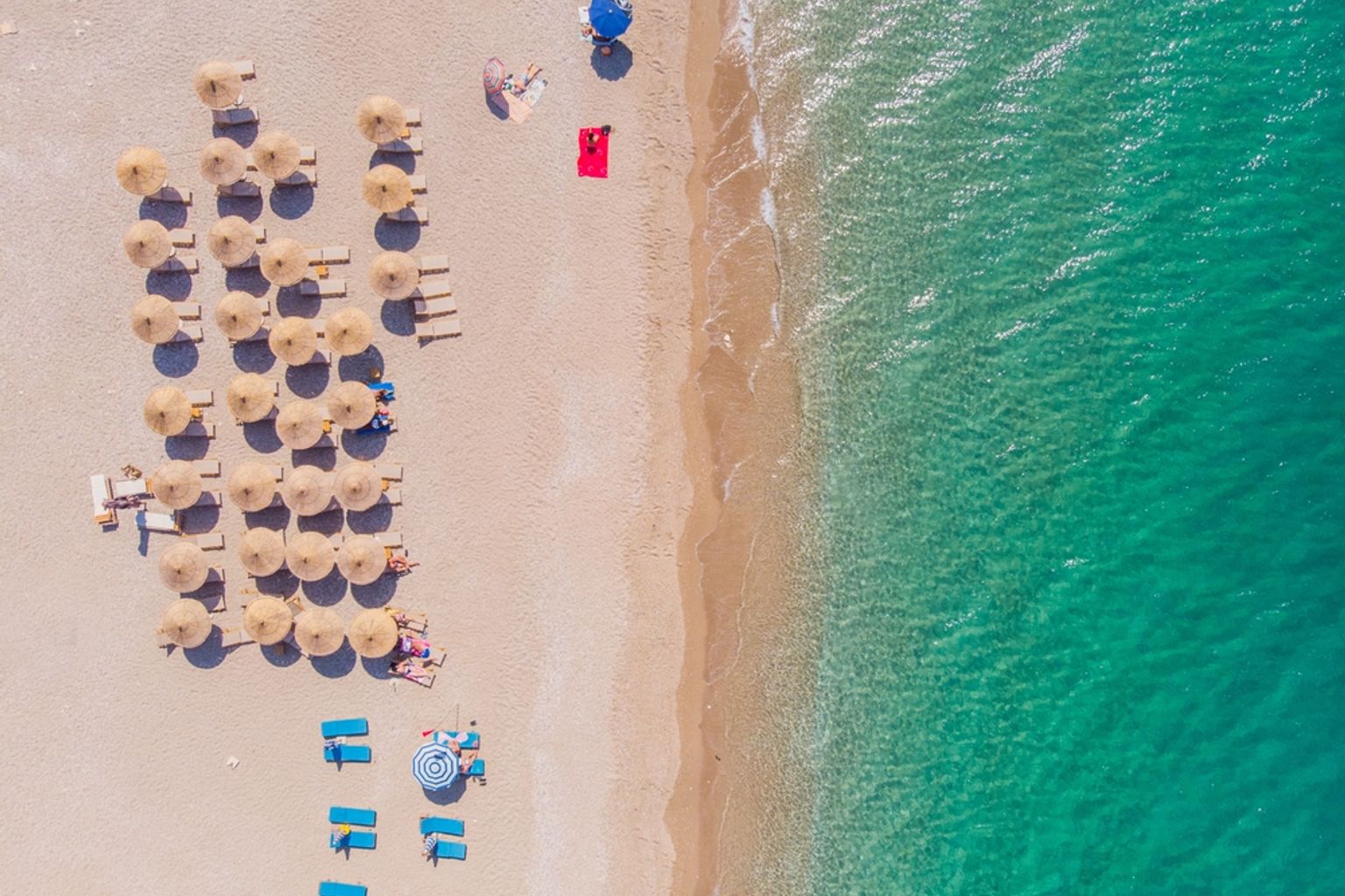 Luftfoto av en sandstrand med rekker av parasoller og solsenger ved siden av klart turkis vann.