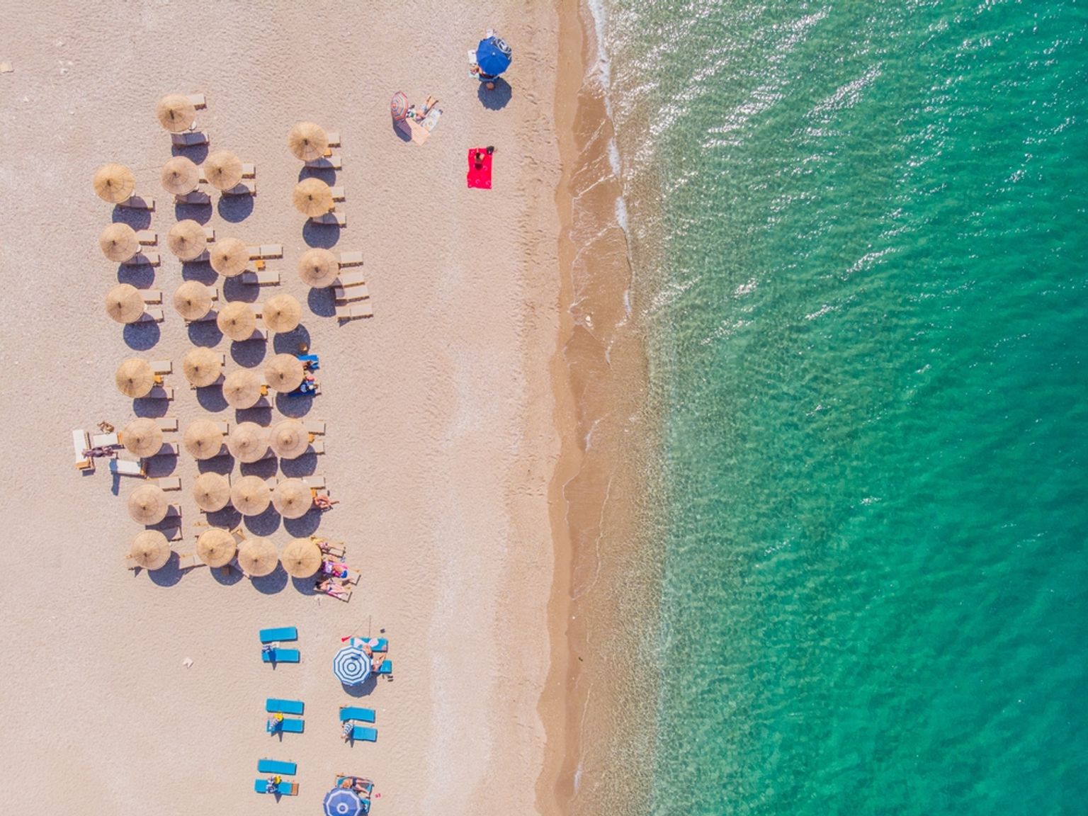 Luftfoto av en sandstrand med rekker av parasoller og solsenger ved siden av klart turkis vann.