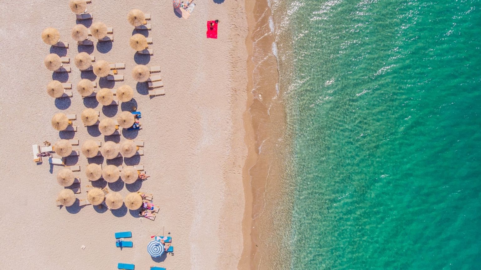 Luftfoto av en sandstrand med rekker av parasoller og solsenger ved siden av klart turkis vann.