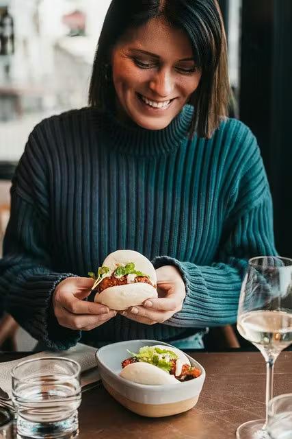 En smilende kvinde i en turkis sweater holder en bao-bolle, med en anden på en tallerken på bordet.