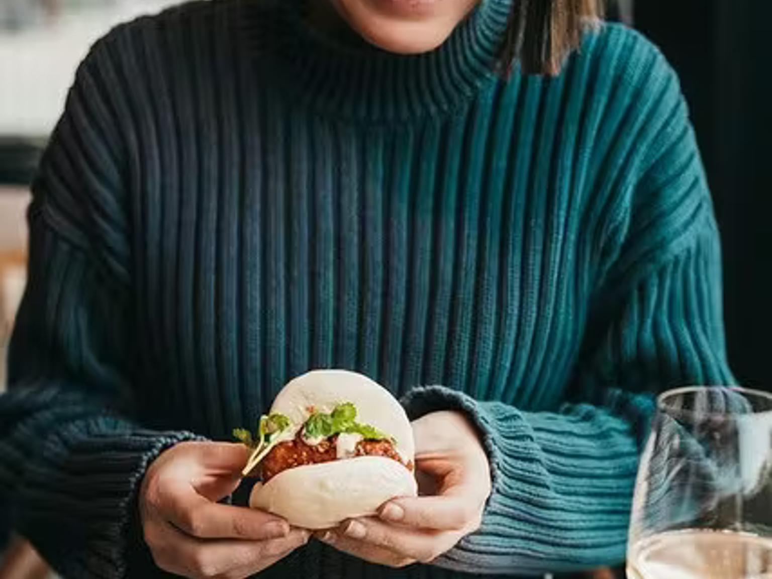 En smilende kvinde i en turkis sweater holder en bao-bolle, med en anden på en tallerken på bordet.