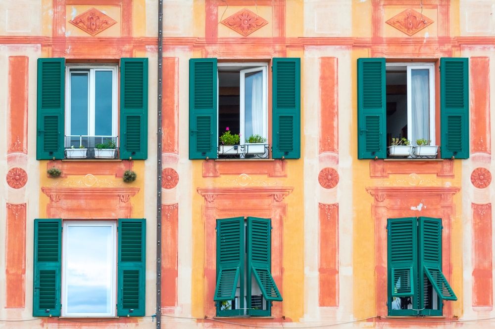 Farverig bygningsfacade med orange og gule striber, med seks vinduer med mørkegrønne skodder, nogle åbne med altankasser. Santa Margherita Ligure.