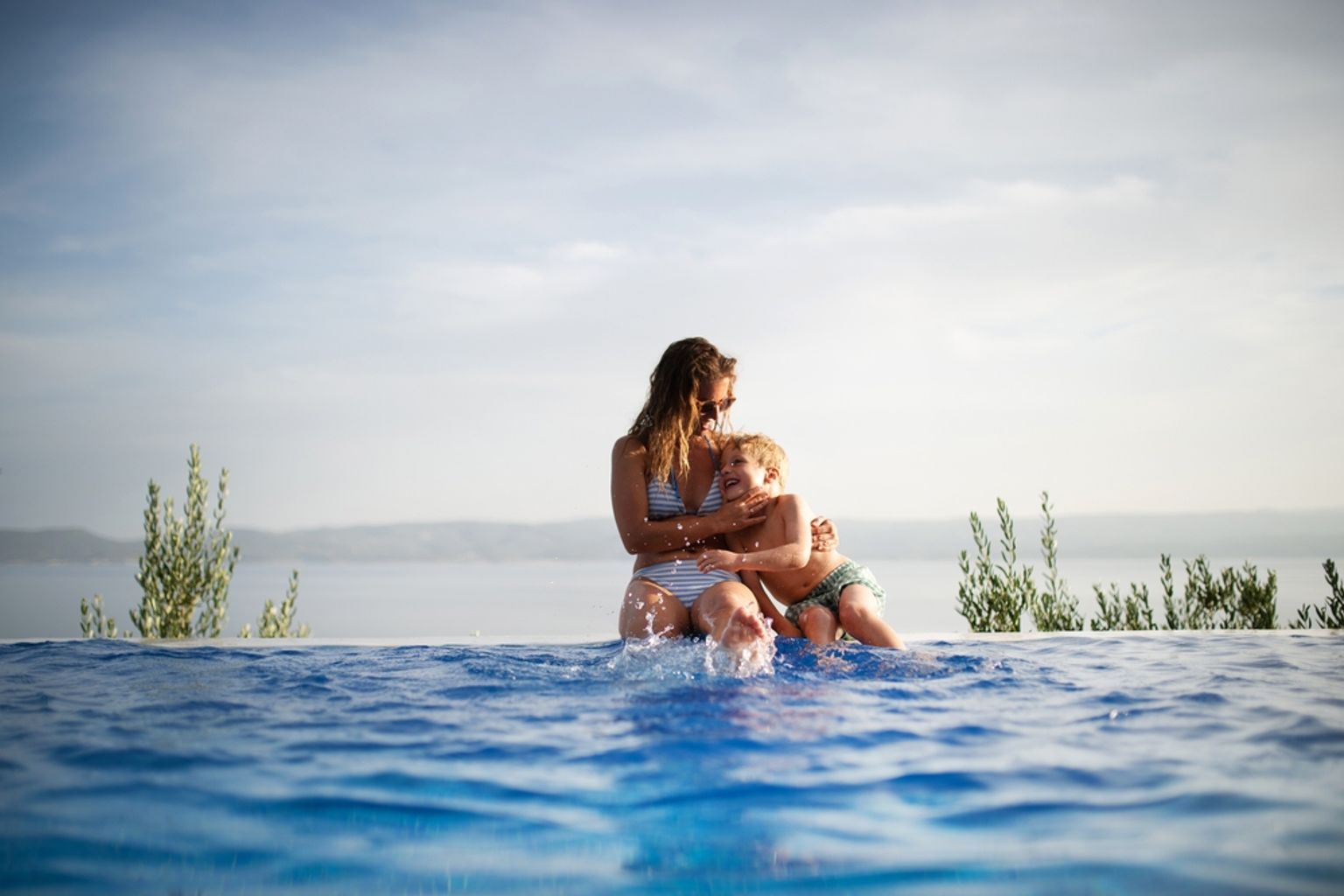 En kvinna och ett barn skvätter vatten vid kanten av en infinitypool med en naturskön havsutsikt.