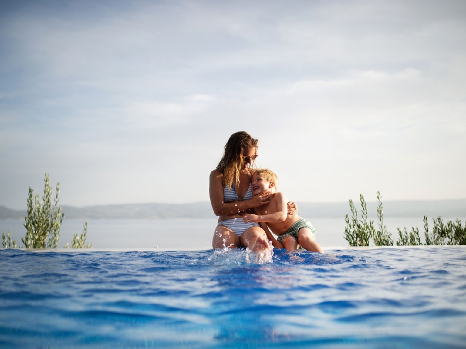 En kvinna och ett barn skvätter vatten vid kanten av en infinitypool med en naturskön havsutsikt.