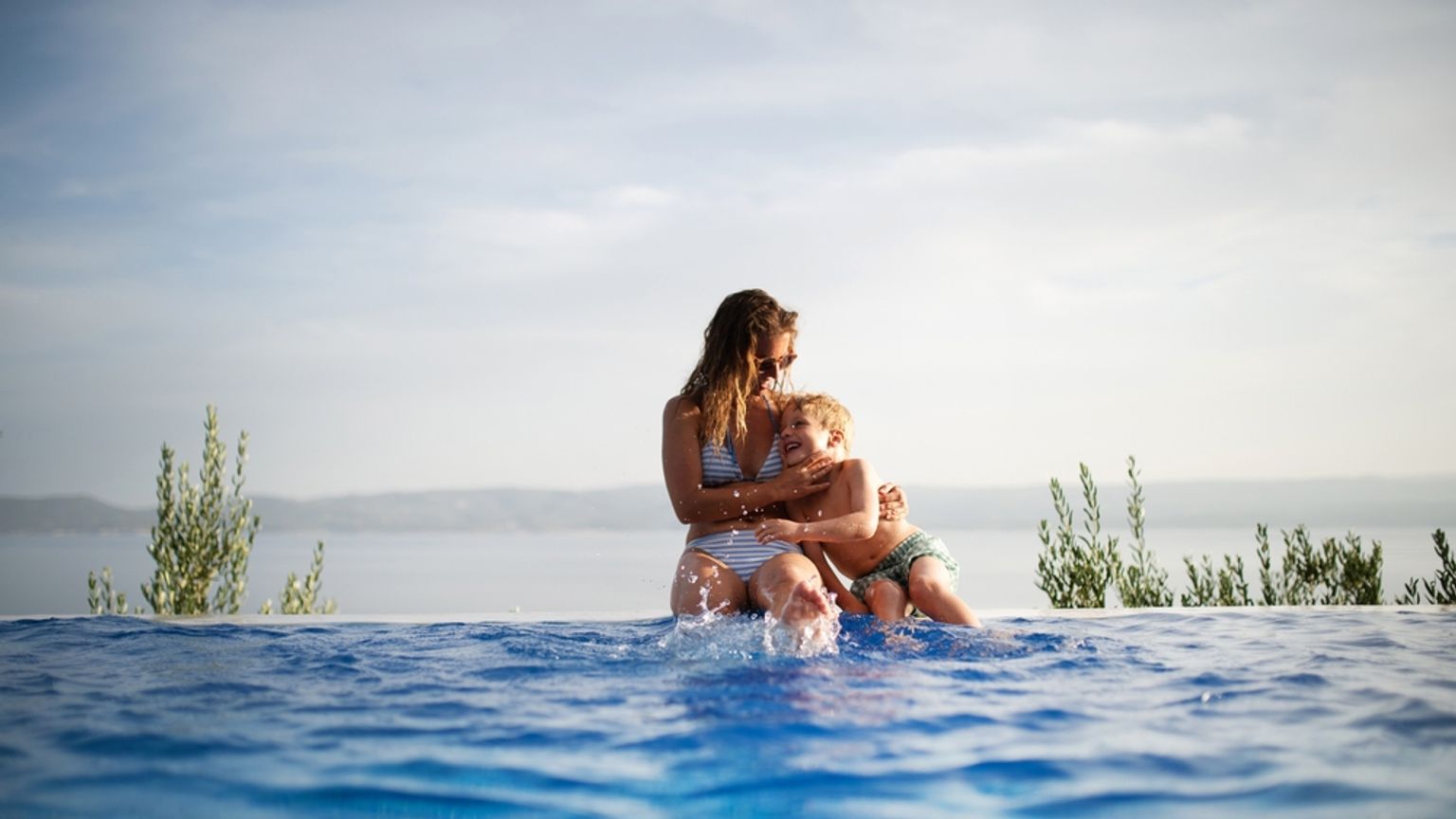 En kvinna och ett barn skvätter vatten vid kanten av en infinitypool med en naturskön havsutsikt.