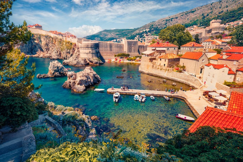 Udsigt oppefra over Dubrovnik i Kroatien med gamle bymure, rødtagede bygninger, en klar turkis bugt med både og klippefremspring.
