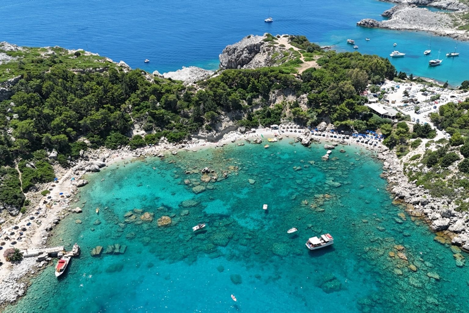 Luftfoto af en livlig turkis bugt med klippefyldte, skovklædte kyster, en strand med parasoller og flere både. Anthony Quinn Bay, Rhodos
