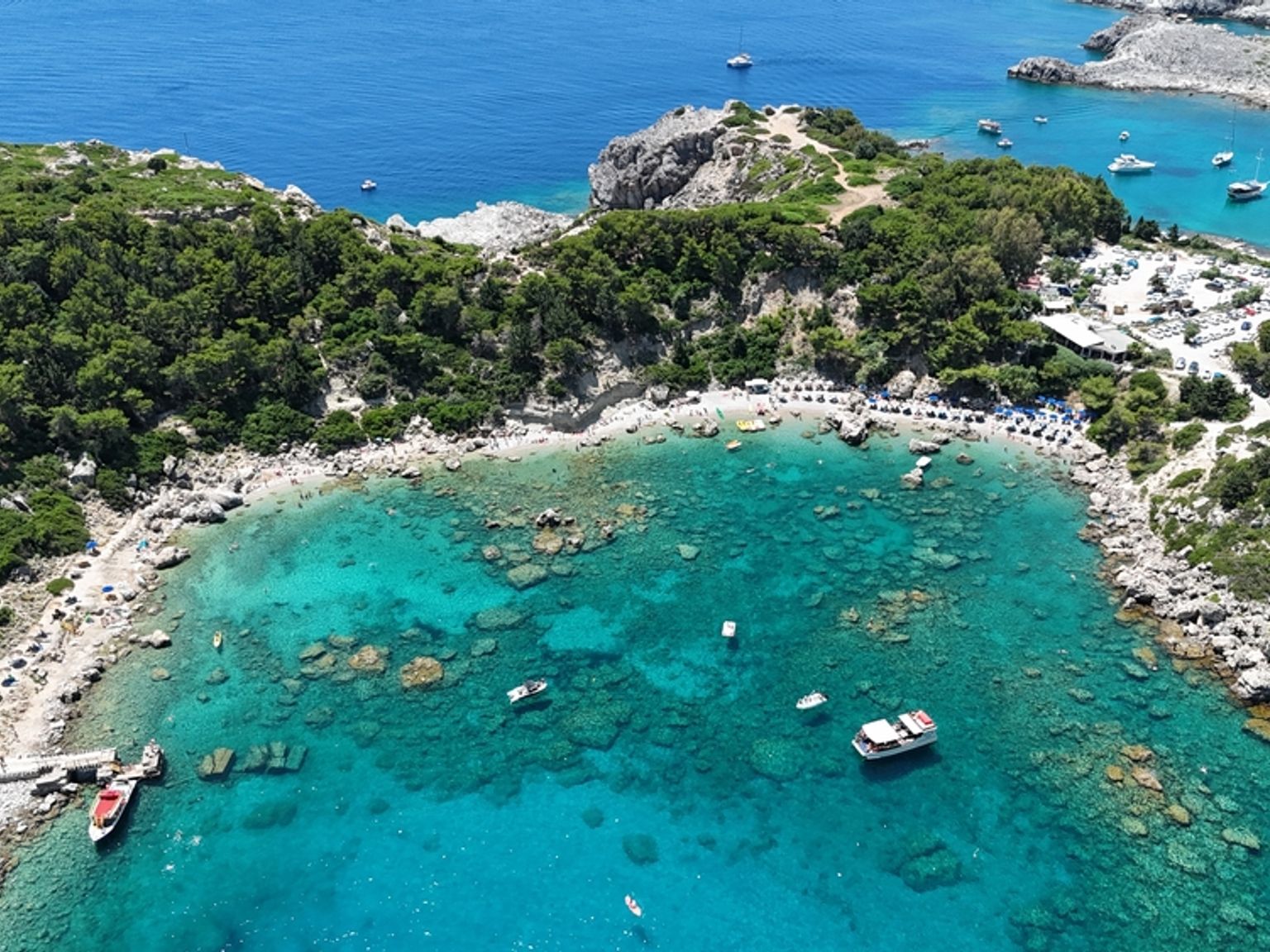 Luftfoto af en livlig turkis bugt med klippefyldte, skovklædte kyster, en strand med parasoller og flere både. Anthony Quinn Bay, Rhodos