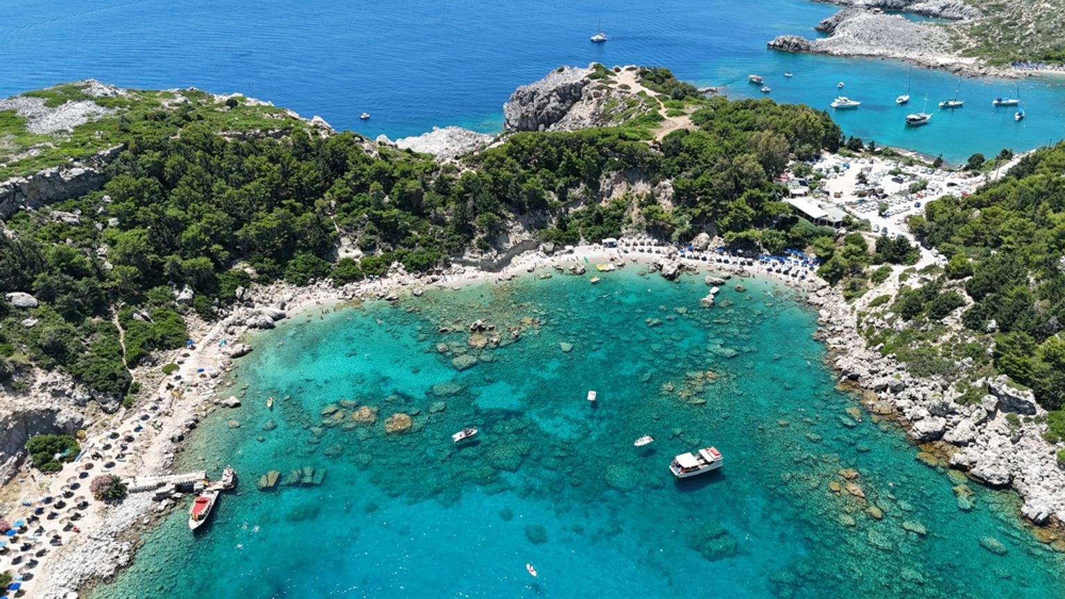 Luftfoto af en livlig turkis bugt med klippefyldte, skovklædte kyster, en strand med parasoller og flere både. Anthony Quinn Bay, Rhodos