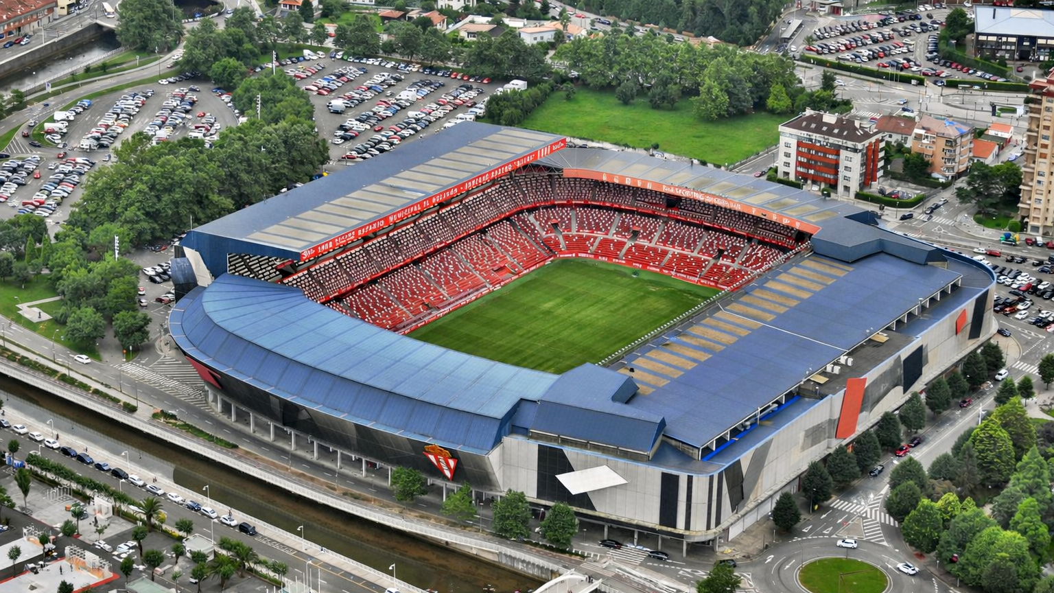 Vista aérea de un estadio deportivo moderno con techos azules, asientos rojos y un campo verde, rodeado por una zona urbana con carreteras, edificios y un río.
