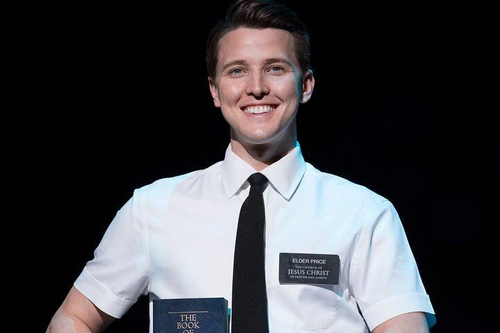 En leende ung man i vit skjorta och svart slips, som bär en namnbricka med texten "Elder Price" och håller i en bok med titeln "The Book of Mormon".